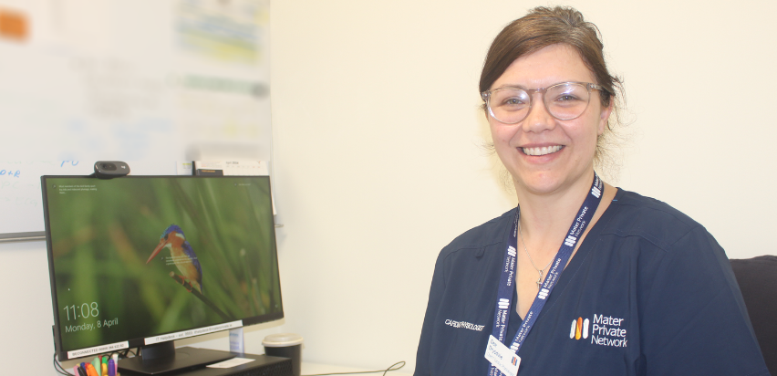 Dee Drysdale is smiling and sitting next to her computer which has a picture of a bird on its screen. She has brown hair and is wearing a blue top.