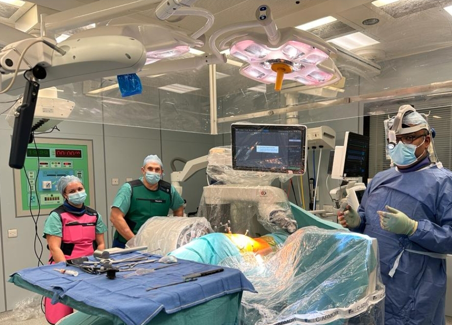Mr. Deb Roy is standing to the right of the image in an operating theatre and is wearing surgical clothing, gloves and a mask. Beside him is a patient who is receiving surgery with the use of a surgical robot which is positioned over the patient.