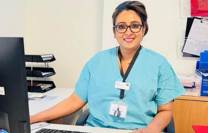 Jincy Kurakose is smiling while sitting in front of a computer screen and keyboard. She is wearing glasses, a green top and a lanyard around her neck.