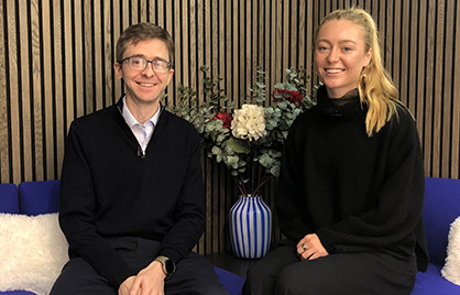 Shane O'Hanlon and Lara Gillespie sitting side by side on blue seats in front of a vase with flowers in it.