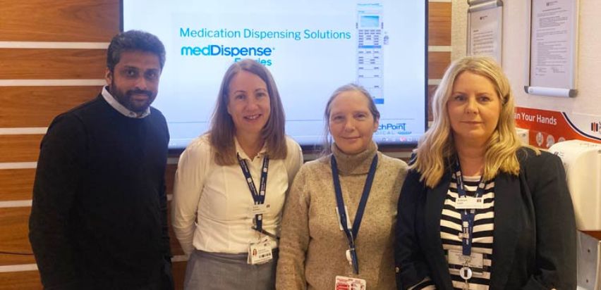 Three female and one male member of the teams involved in introducing the new medication equipment are standing together in front of a screen which displays the MedDispense logo.