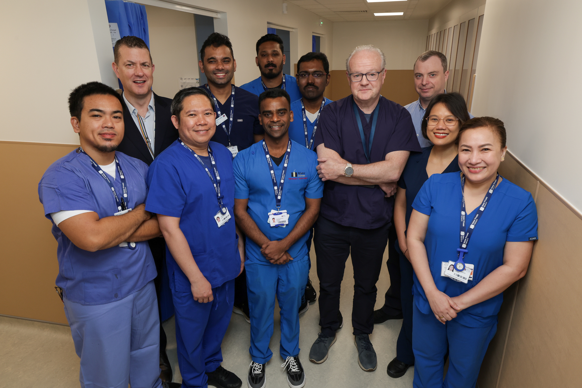 The Emergency Department team are standing side by side. They are wearing a mixture of light blue and dark blue scrubs and are posing beside the new patient bays.