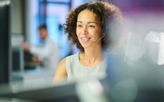 A woman is sitting at a desk and is looking at a computer while speaking through a headset.