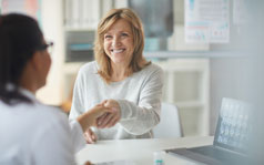 A woman is smiling and shaking the hand of a female doctor sitting across from her.