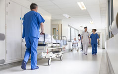 Various medical professionals walking down the corridor of a hospital. One man is wheeling a patient bed.