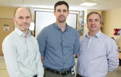Three of the clinical engineers, Tom Bergin, Gary McDonnell and Glenn Tunney stand side by side in front of a large pulldown screen. Tom and Glen are wearing bright coloured shirts while Gary who is in the middle is wearing a dark blue checkered shirt.