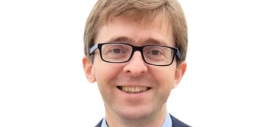 A man with light brown hair in a suit wearing glasses and smiling