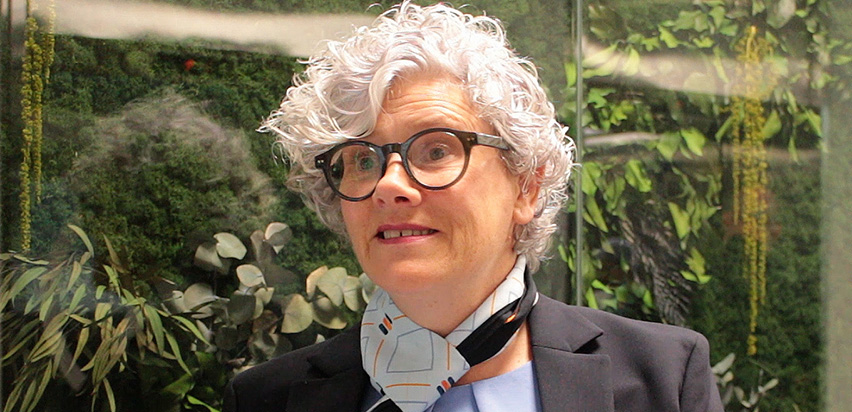 Zita McGarry Kelly is smiling and standing in front of a wall decorated with flowers and leaves.