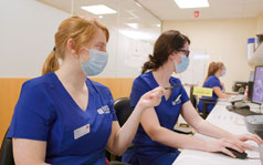 Two medical professionals wearing medical masks are looking at a computer screen.