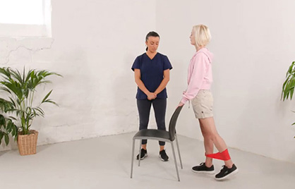 A physiotherapist working with a patient who is doing exercises with a chair and a red band on her legs.